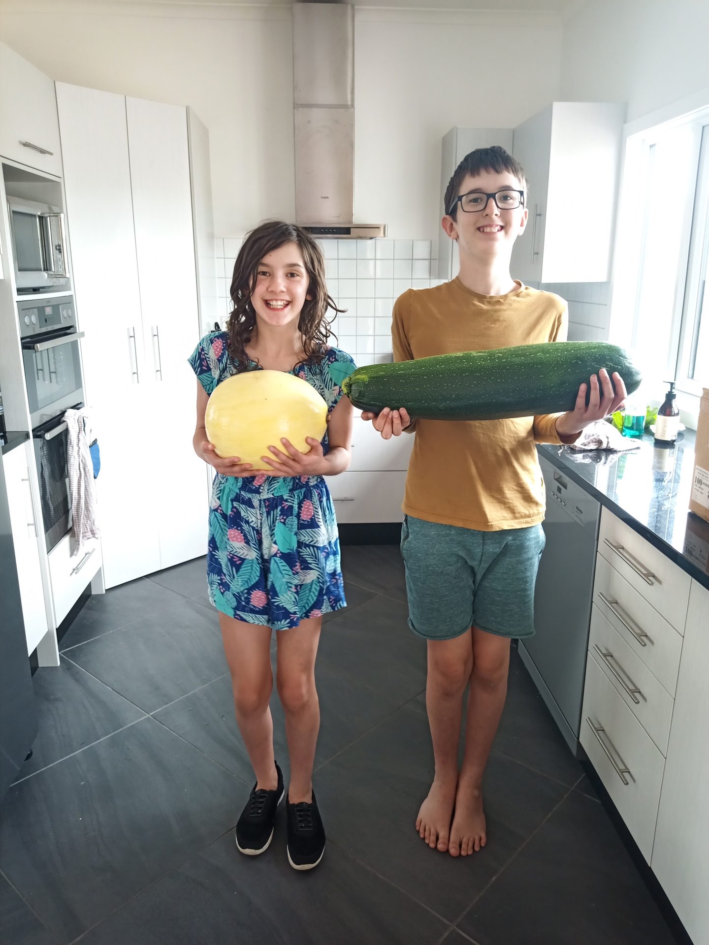 Two children in a kitchen holding up an enormous marrow and a large melon grown on the property