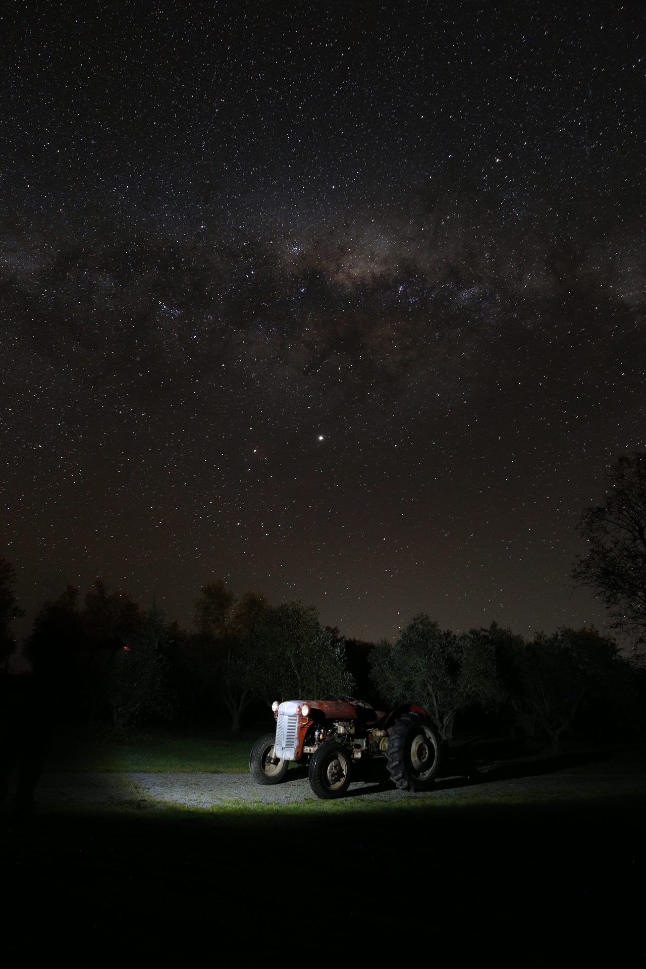 The Milky Way arching from horizon to horizon above a lit Ferguson tractor