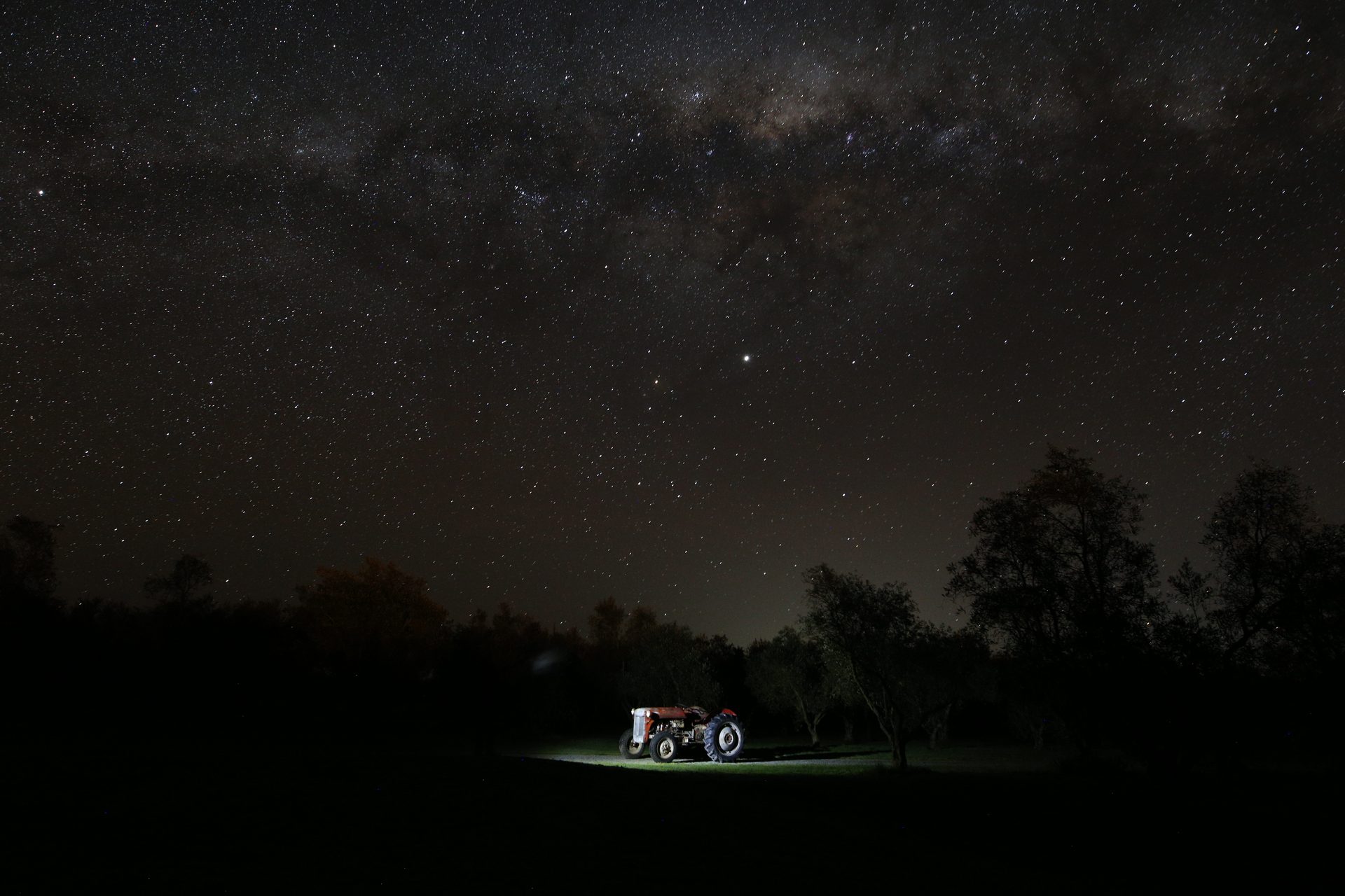 The Milky Way arching over a vintage Ferguson tractor lit by torchlight in a mown olive grove at night