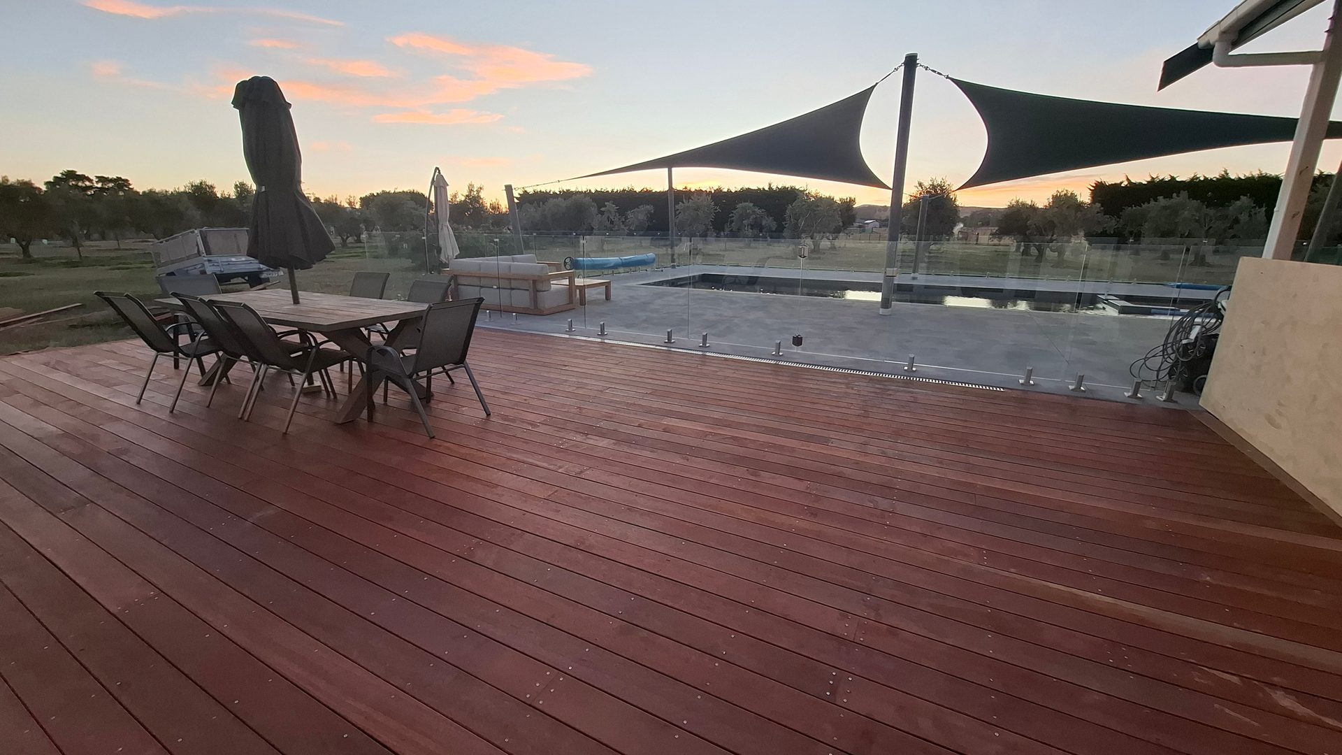 A large hardwood deck with outdoor dining table, shade sails and a swimming pool at dawn