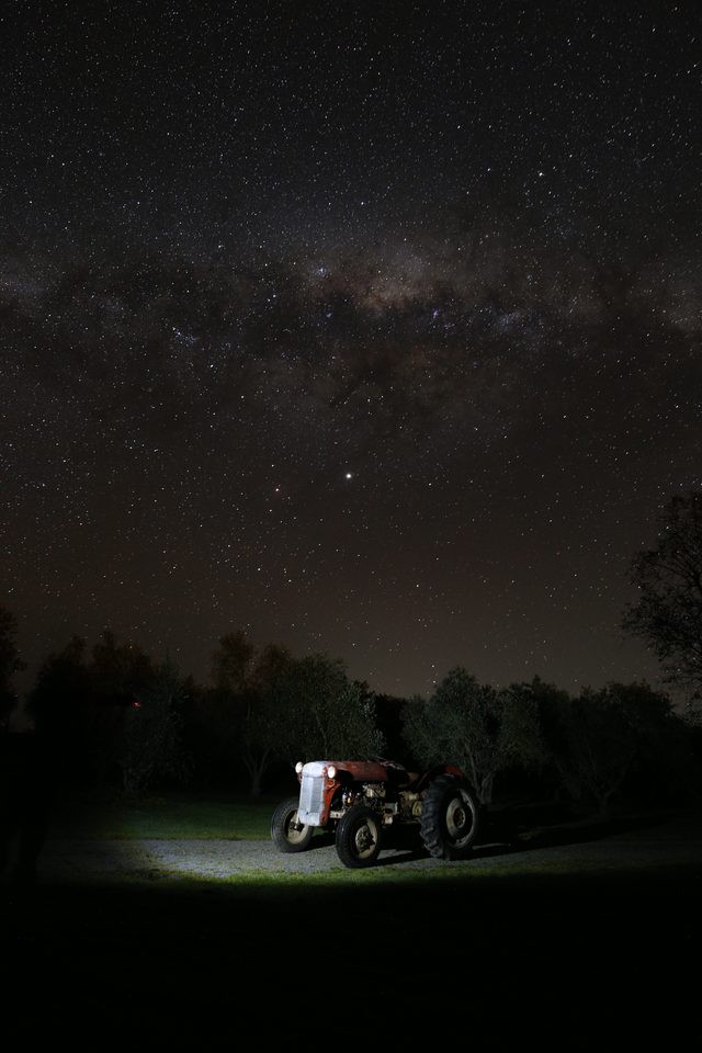 The full arch of the Milky Way above a lit tractor in the grove