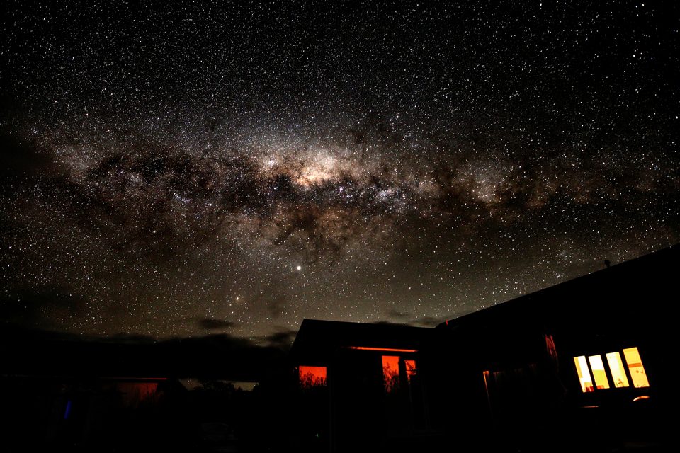 The bright core of the Milky Way above the house, its windows glowing warm orange